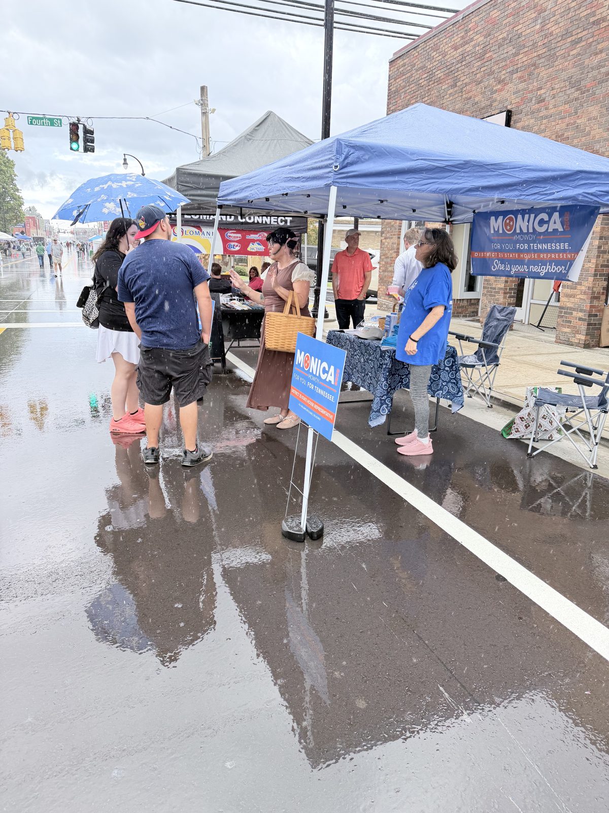 Monica campaign tent on a rainy day at a downtown street festival, with a volunteer talking to visitors
